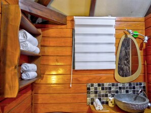 A wooden bathroom with a shelf holding rolled towels, a mirror, a sink with a stone basin, and a tiled backsplash.