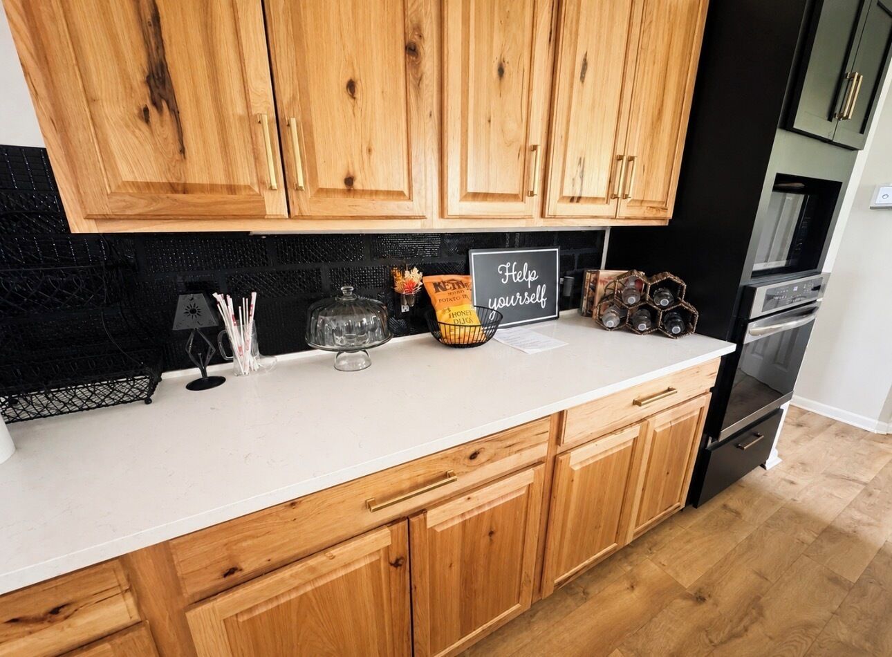 A kitchen with wooden cabinets, a white countertop, and a black backsplash. There is a sign that says 'Help yourself' and a basket of potatoes.