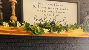 A cozy seating area with a fireplace, a decorative sign above, and a wooden shelf with greenery.