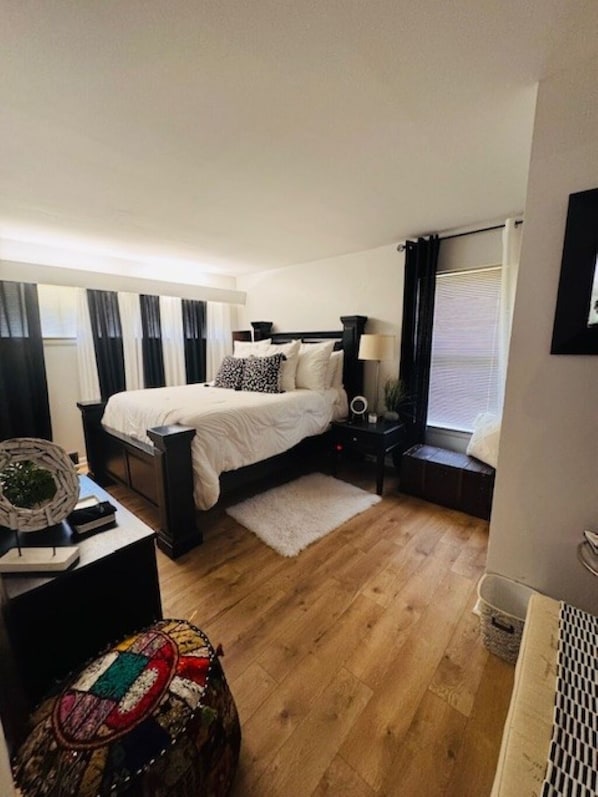 Une chambre à coucher avec un lit, des tables de chevet, une lampe, une fenêtre avec des rideaux, un tapis et un petit tabouret.