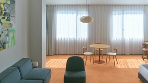 Quarto de hotel com sofá, mesa de jantar com cadeiras, cama e uma janela grande com cortinas.