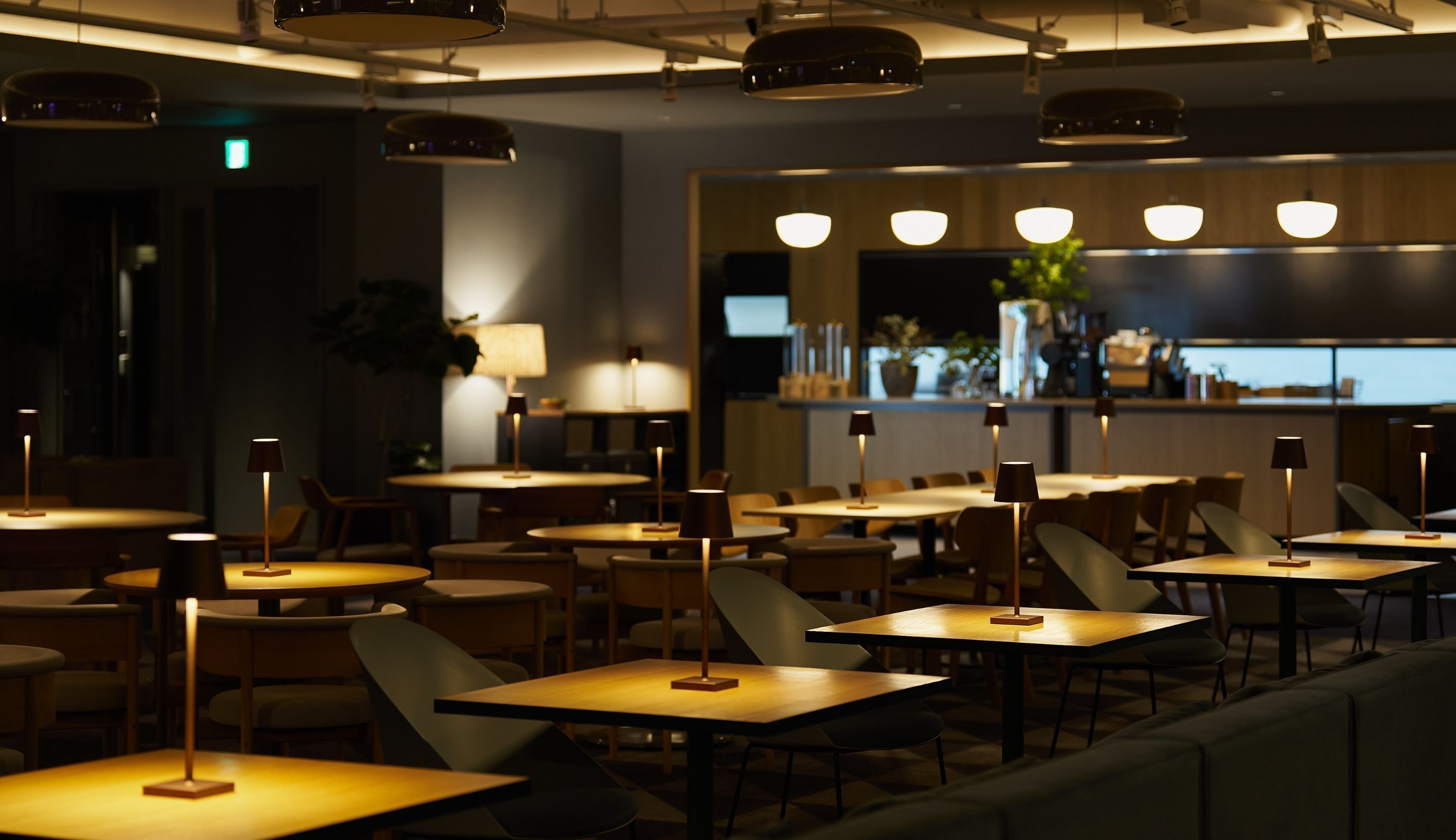 Ein Restaurantinneres mit mehreren Tischen und Stühlen, Deckenleuchten und einer Bar im Hintergrund.