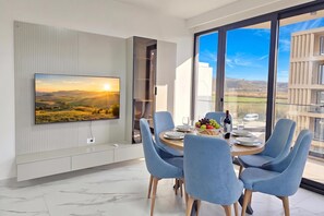 A modern dining area with a large TV mounted on the wall, a round table set for four, and a scenic view through large windows. - Pool Side Apartment, Vlore, Albania (Vlore)
