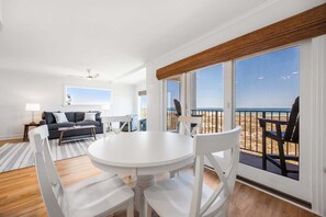 Un salon avec une table et des chaises blanches, un canapé et une vue sur la plage grâce à de grandes fen êtres.