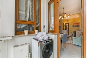 A laundry area with a washing machine, a dining table with chairs, and a sofa in a room with wooden windows and a chandelier. - La Torinese, Torino, Italy (Torino)
