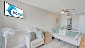 Un saló amb un sofà blanc, un ventilador de peu i una televisió de pantalla plana que mostra 'BIG FISH Rentals'.