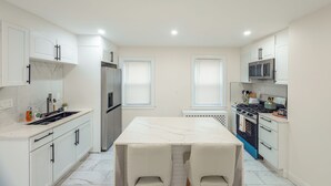 A modern kitchen with white cabinetry, a marble countertop, a stainless steel refrigerator, a stove, and two white chairs.