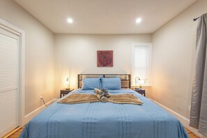 A bedroom with a bed, two bedside tables, a nightstand, a wall-mounted artwork, and a window with blinds. - Serene 3BR-1BA Apt in vibrant Boston - Near T w Parking (Boston)