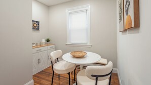 A dining area with a round table, four chairs, a window with blinds, a cabinet with drawers, and a small plant on the counter.