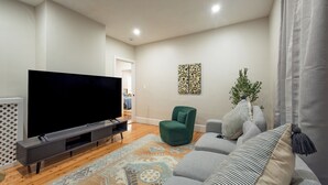 A living room with a flat-screen TV, a green armchair, a patterned rug, and a grey sofa.