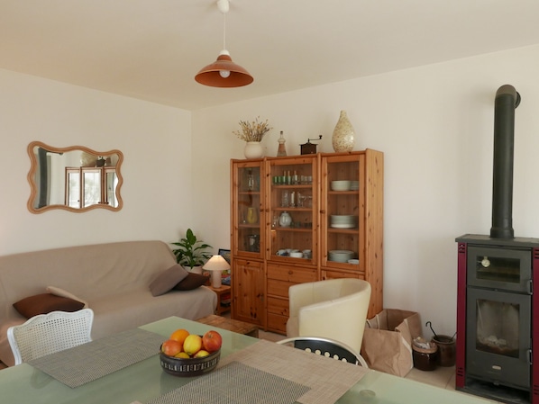 Una sala de estar con un sofá, una mesa de comedor con un frutero, un aparador con cristalería y un estufa de leña.