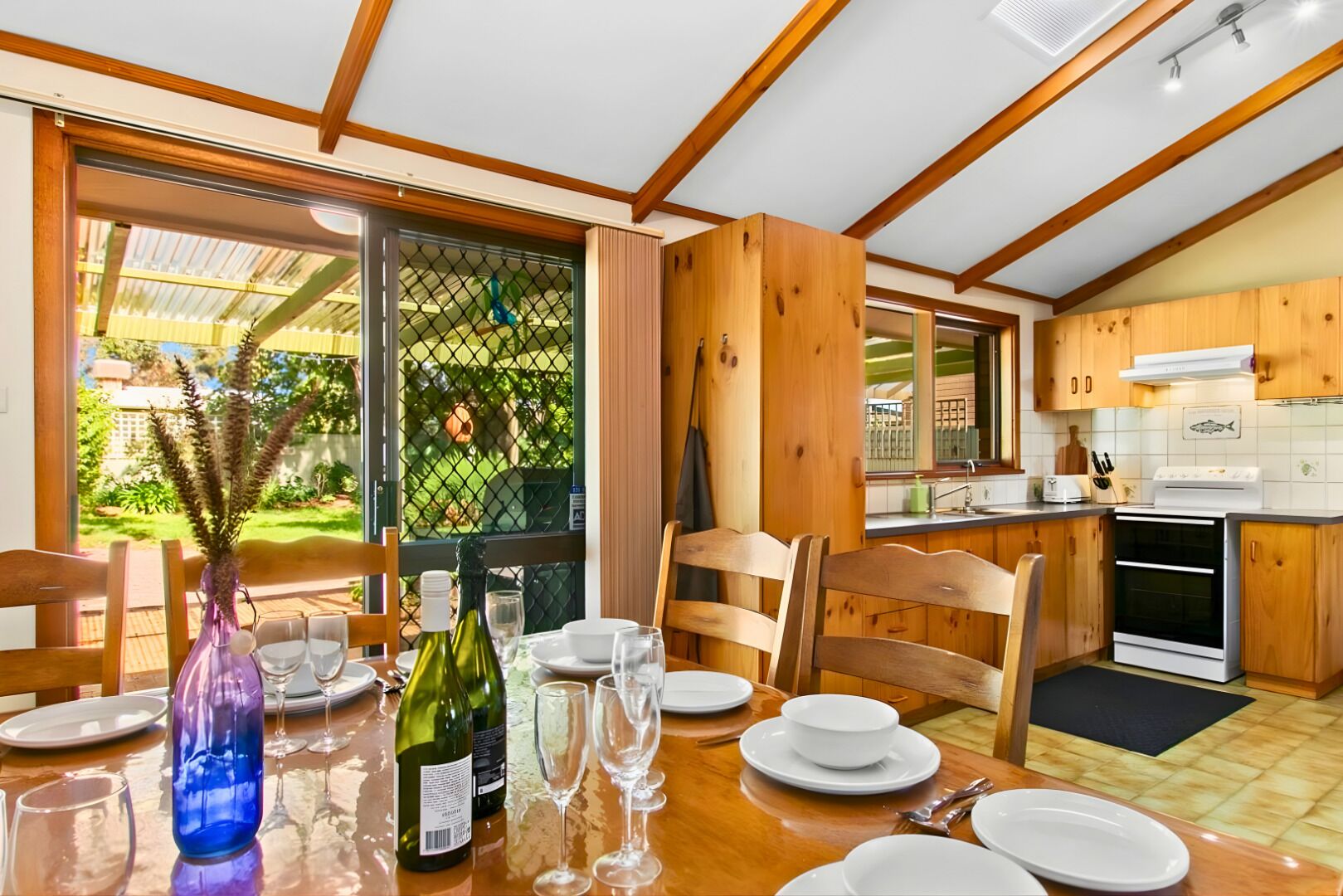 Un comedor con una mesa dispuesta para cuatro, una cocina con armarios de madera y vistas a un jardín por una puerta de cristal.