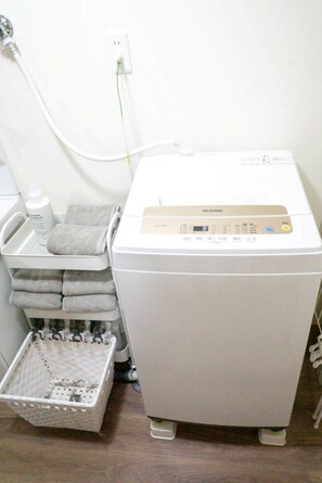 Una lavadora blanca con panel de control, una cesta de toallas dobladas y una botella de detergente líquido.