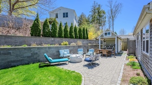 Ein Garten mit einer Steinterrasse, Sitzgelegenheiten im Freien, einem kleinen Schuppen und einem Haus im Hintergrund.