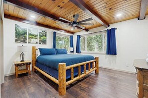 A bedroom with a wooden bed, a ceiling fan, large windows with curtains, and artwork on the wall.