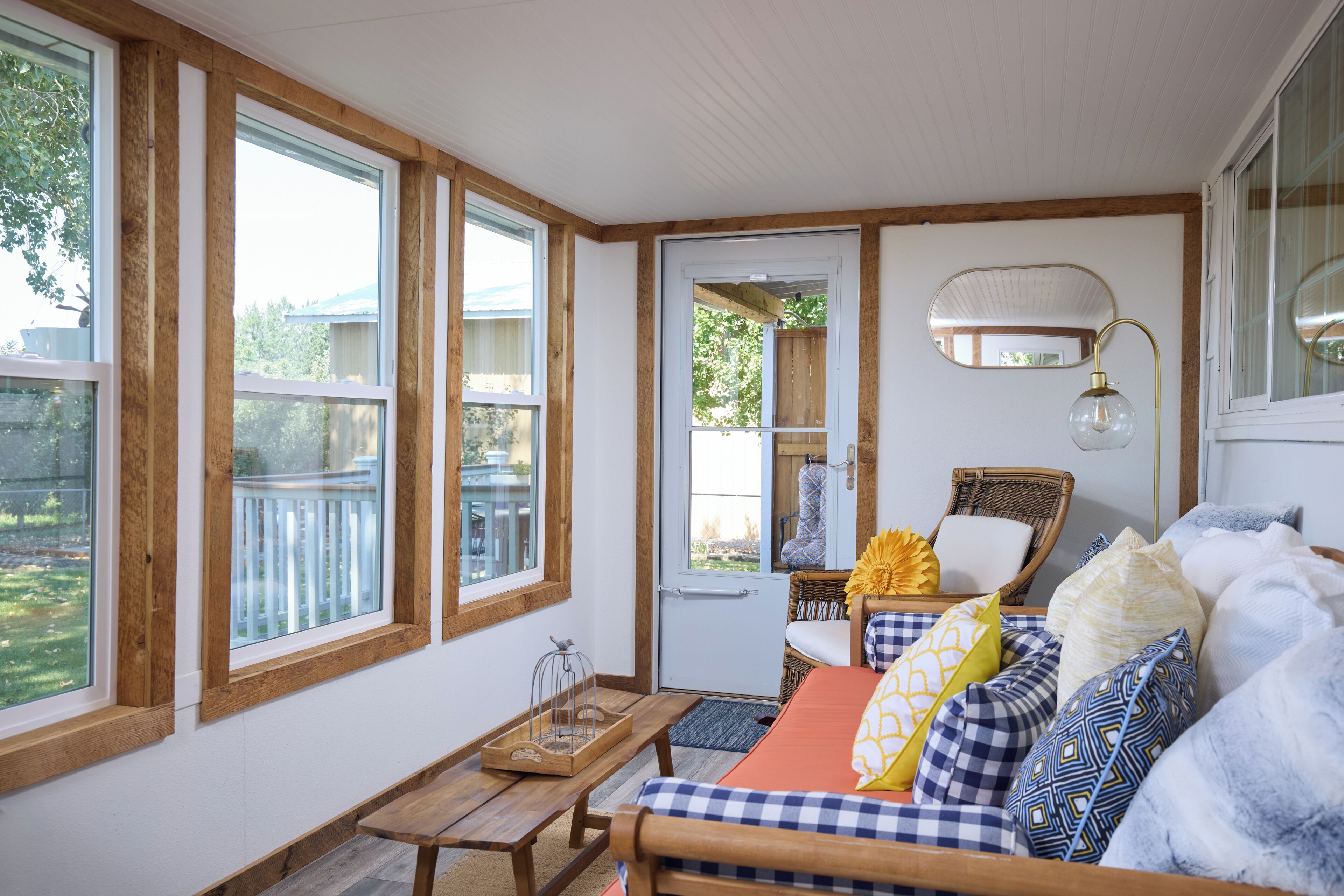 A cozy room with large windows, a wicker chair, a wooden bench, and a bed with patterned pillows.