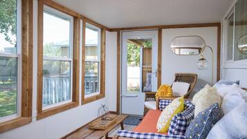 A cozy room with large windows, a wicker chair, a wooden bench, and a bed with patterned pillows.