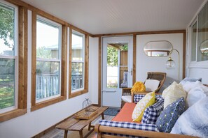 A cozy room with large windows, a wicker chair, a wooden bench, and a bed with patterned pillows.