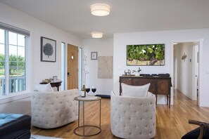 A spacious living room with two upholstered chairs, a small round table with wine glasses, a wooden cabinet, and a large flat-screen TV mounted on the wall. - Sleep Inside Wine Country | 4BR, Fireplace (Milton-Freewater)