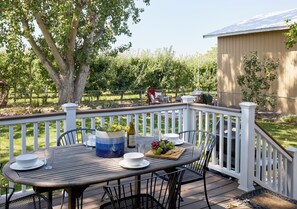 An outdoor deck with a table set for dining, surrounded by chairs, and a large tree in the background. - Sleep Inside Wine Country | 4BR, Fireplace (Milton-Freewater)