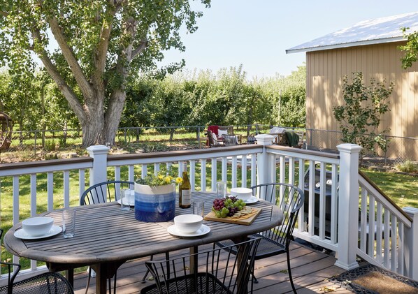 An outdoor deck with a table set for dining, surrounded by chairs, and a large tree in the background.