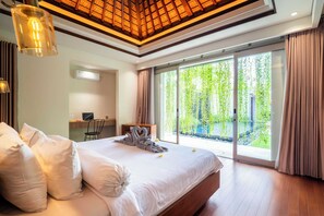 A modern hotel room with a large bed, a desk, and a view of greenery through the sliding glass door. - Bali KBS Villas at Jimbaran Bay 1 BR (Badung)