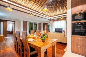 A dining area with a wooden table, chairs, and a vase of flowers. There is a kitchen area with wooden cabinets and a built-in oven. - Bali KBS Villas at Jimbaran Bay 1 BR (Badung)