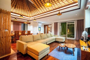 A modern living room with a sectional sofa, wooden flooring, a dining area with a table and chairs, and a large sliding glass door leading to a balcony. - Bali KBS Villas at Jimbaran Bay 1 BR (Badung)
