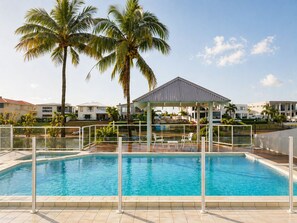 A swimming pool with a covered seating area, palm trees, and modern buildings in the background. - Sip, Swim, Repeat - 5BR Waterfront Pool Escape (Mermaid Waters)