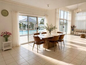 A spacious dining area with a large table and chairs, a view of a swimming pool, and a sofa in the background. - Sip, Swim, Repeat - 5BR Waterfront Pool Escape (Mermaid Waters)