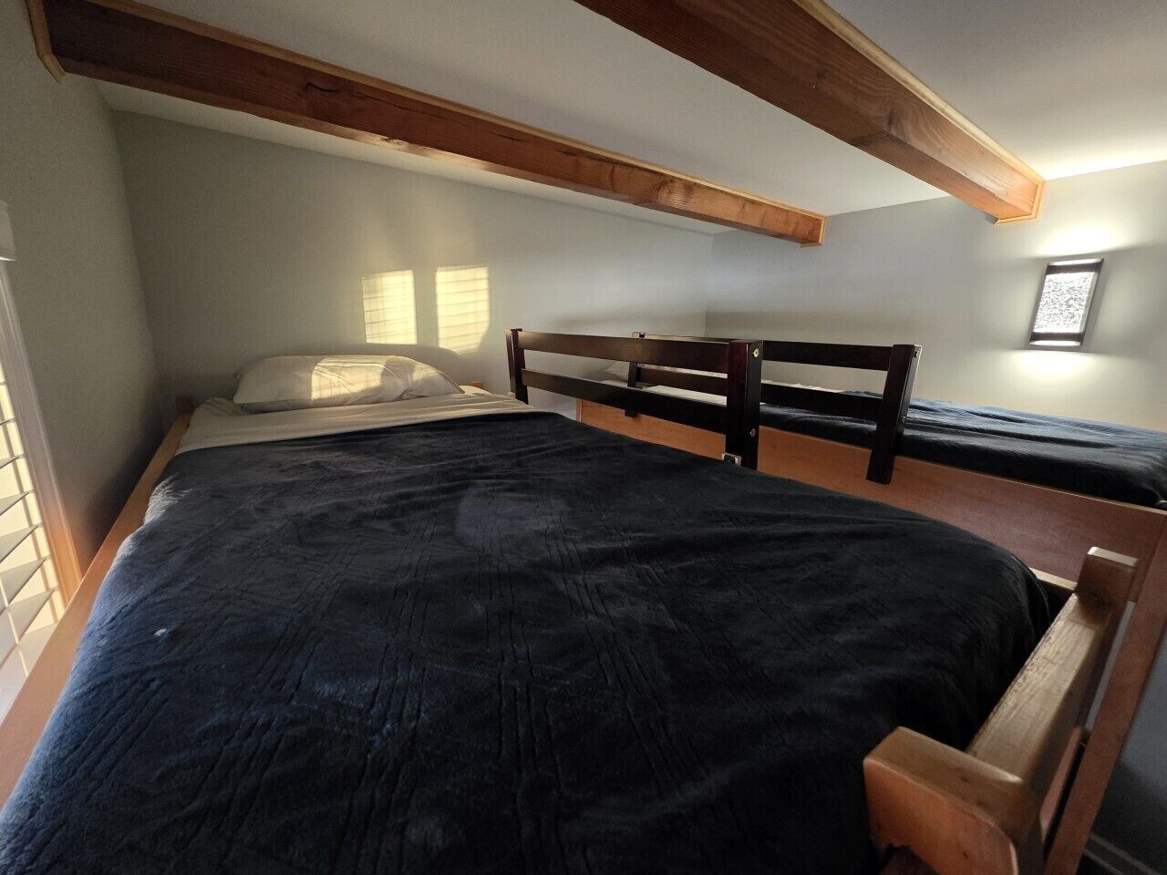 A single bed with a wooden headboard, a dark bedspread, and a window with blinds.