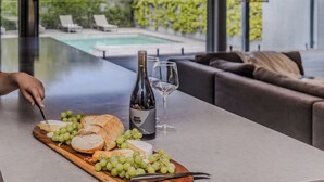 A table with cheese, grapes, a bottle of wine, and a glass, with a pool and greenery visible through large windows. - Thule Country Estate | Sleeps 12 | Private Lake, Pool & Farm | 45 mins Melbourne (Mount Macedon)