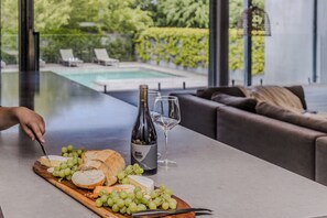 A table with cheese, grapes, a bottle of wine, and a glass, with a pool and greenery visible through large windows. - Thule Country Estate | Sleeps 12 | Private Lake, Pool & Farm | 45 mins Melbourne (Mount Macedon)