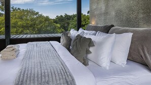 A bed with a view of trees through a window, a concrete wall, and a grey blanket. - Thule Country Estate | Sleeps 12 | Private Lake, Pool & Farm | 45 mins Melbourne (Mount Macedon)