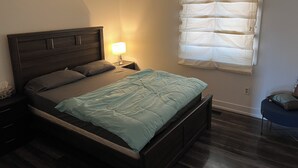 A bedroom with a bed, nightstand, lamp, window with blinds, and a chair. - Cozy Queen Room (Brampton)