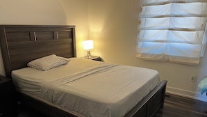 A bedroom with a wooden bed, a nightstand, a bedside lamp, and a window with blinds. - Cozy Queen Room (Brampton)