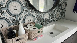 A bathroom with a white sink, a mirror, and patterned tiles. There are two baskets on the counter, one with a bottle and the other with toilet paper. - Cozy Queen Room (Brampton)
