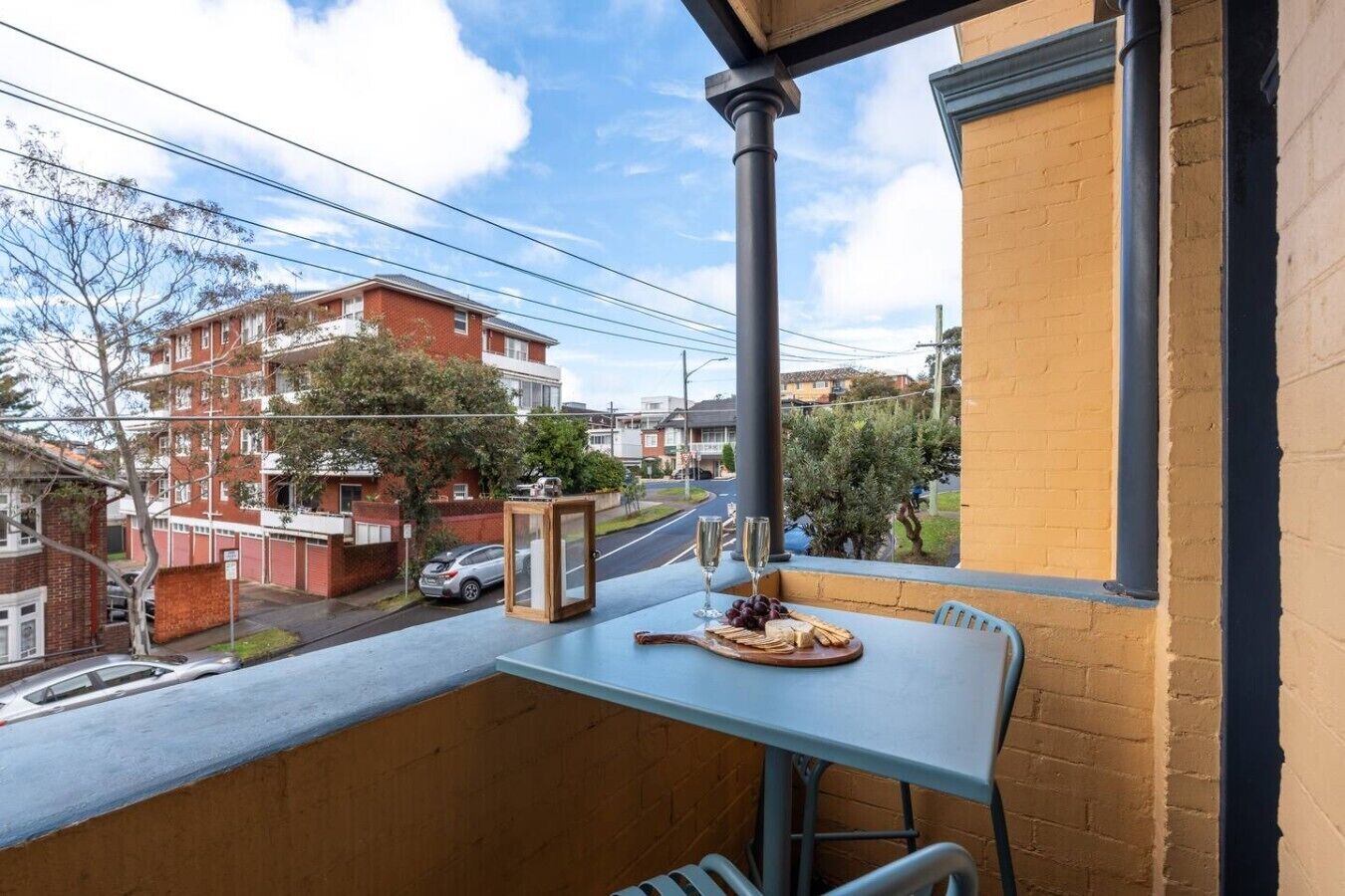 Ein Balkon mit einem Tisch für zwei Personen, mit Blick auf eine Straße mit parkenden Autos und Gebäuden.
