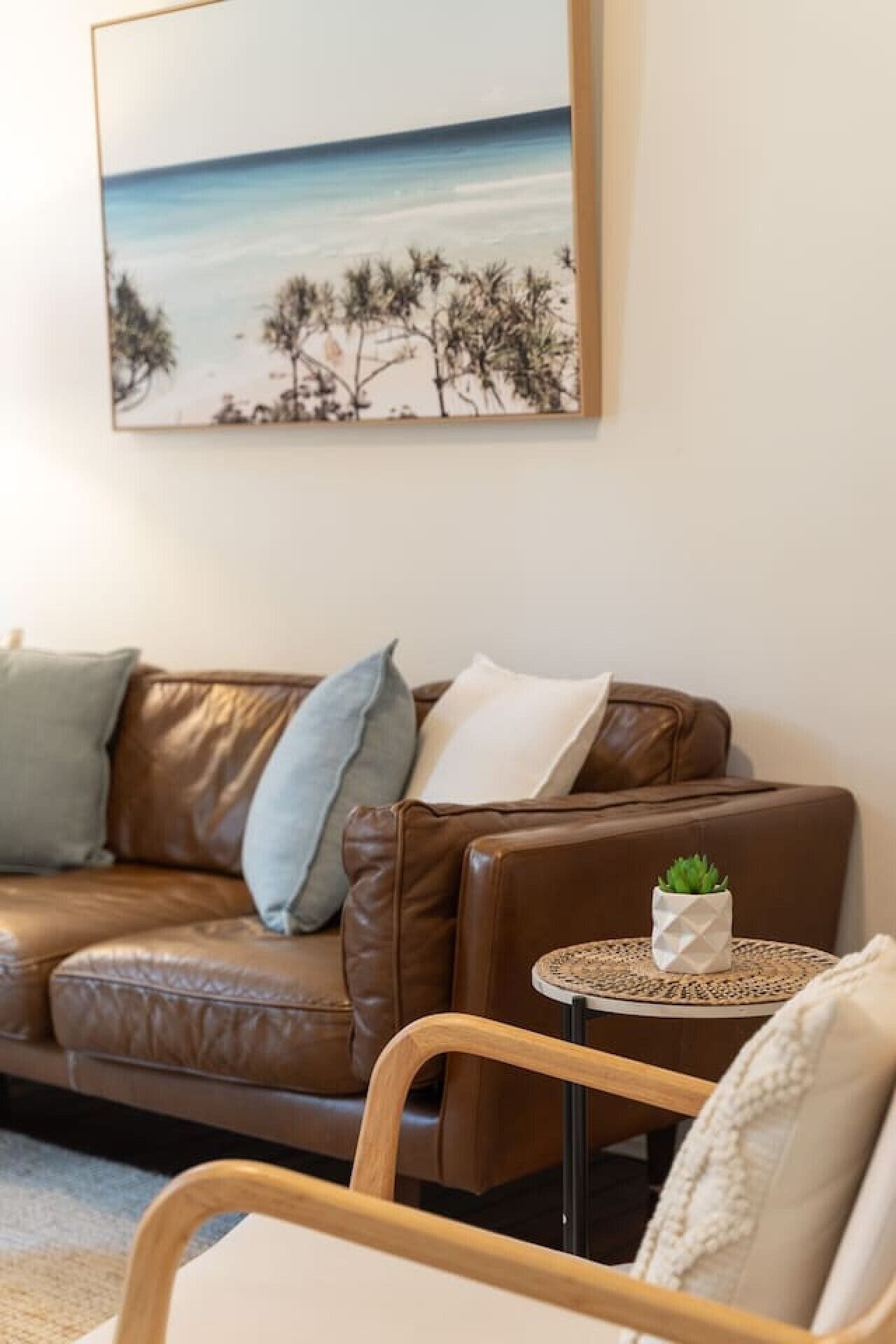 A brown leather sofa with cushions, a small round table with a plant, and a painting of a beach scene on the wall.