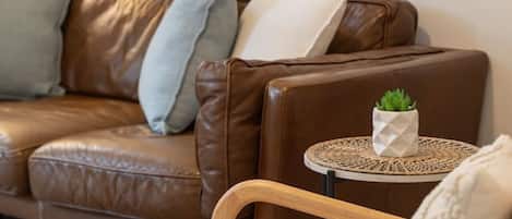 A brown leather sofa with cushions, a small round table with a plant, and a painting of a beach scene on the wall.