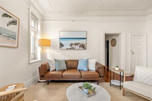 A living room with a brown leather sofa, a round coffee table, and a painting of a beach scene. - Art Deco Coogee Apartment | 5-Min Walk to Beach (Coogee)