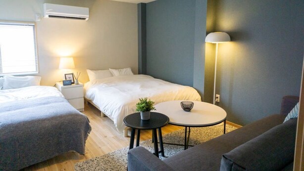 Quarto de hotel moderno com duas camas, uma mesinha redonda, abajur, planta em vaso e janela com persianas.