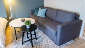 Um quarto com um sofá cinzento, uma mesa redonda com uma planta em vaso, um abajur e uma parede branca.