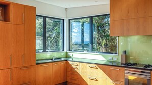 A modern kitchen with wooden cabinets, a stainless steel stove, and large windows offering a view of trees and a body of water. - Hekereua Heights - Spa & Ocean Views (Waiheke Island)