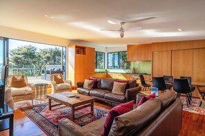 A spacious living area with a large window, a ceiling fan, and a dining area with black chairs. - Hekereua Heights - Spa & Ocean Views (Waiheke Island)