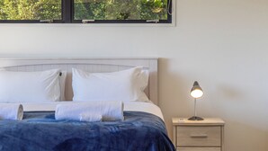 A neatly made bed with white pillows and a blue blanket, a bedside lamp, and a nightstand with drawers. - Hekereua Heights - Spa & Ocean Views (Waiheke Island)