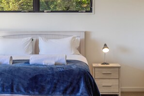 Una cama bien hecha con almohadas blancas y una manta azul, una lámpara de noche y una mesita de noche con cajones.