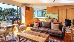 A modern living room with a leather sofa, wooden coffee table, wicker chairs, and a dining area. - Hekereua Heights - Spa & Ocean Views (Waiheke Island)