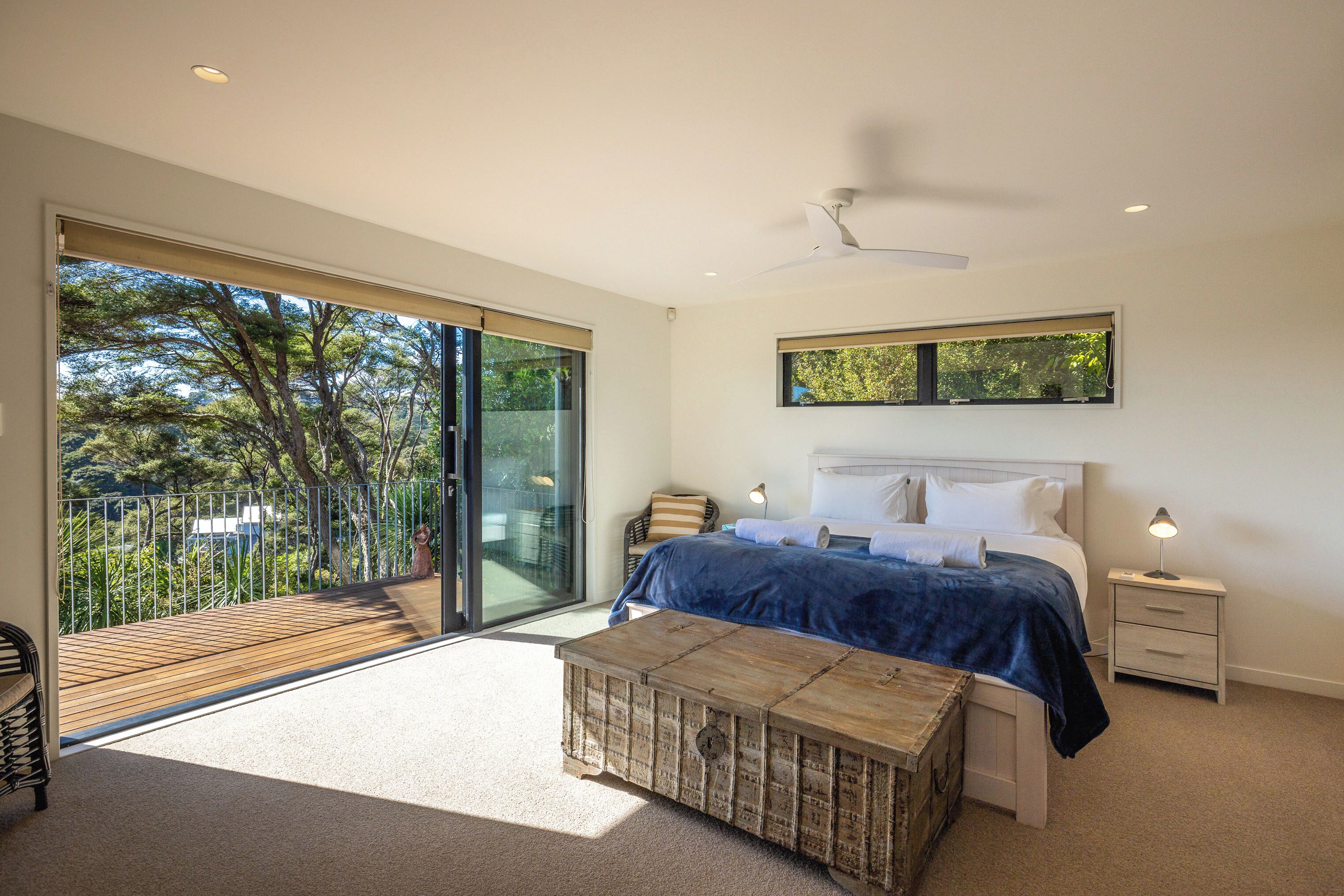A bedroom with a large sliding glass door leading to a balcony, a bed with a blue blanket, a wooden bench, and a ceiling fan.