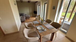 A dining area with a wooden table, four chairs, a television, and a view of the outdoors through large windows. - Stylishly furnished first floor apartment with garden view (Rheinsberg)